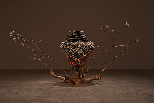 A sculptural installation featuring a central, textured form composed of layered materials, resembling a natural growth. Delicate, branching elements extend outward, creating a sense of balance and tension.